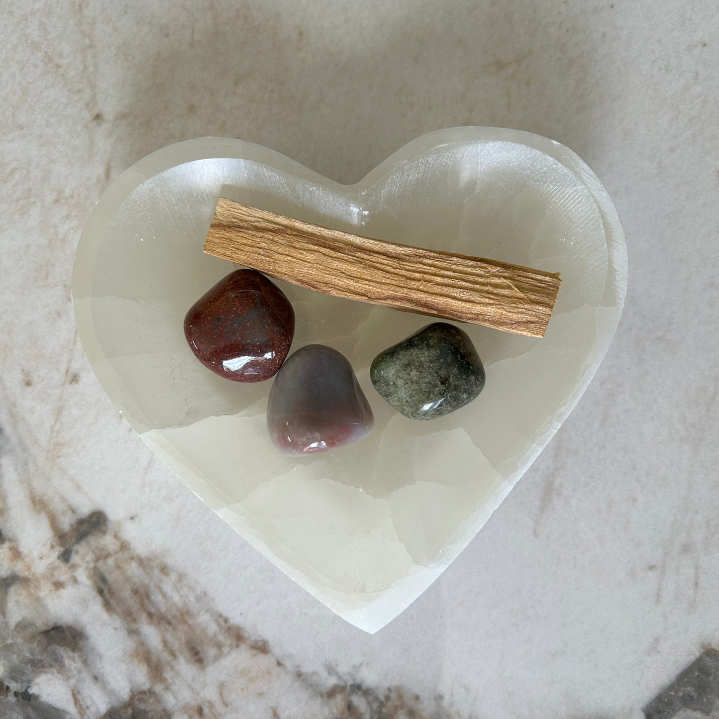 Bol coeur en sélénite posé sur du marbre. Contient trois pierres roulées et un bâtonnet de palo santo