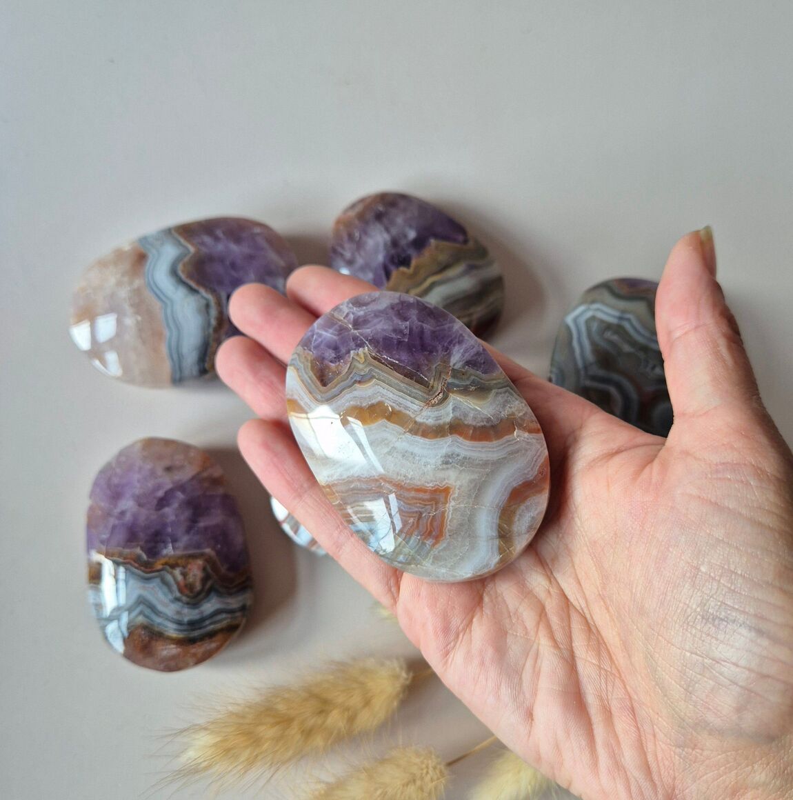 Galet d'agate améthyste posé dans une main avec en fond quatre autres galets et des herbes séchées