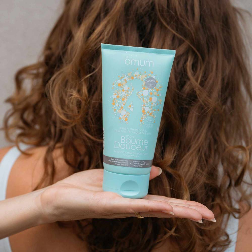 Main de femme tenant un tube bleu lagon de l'après shampoing Mon Baume Douceur Omum, devant une chevelure de femme de dos