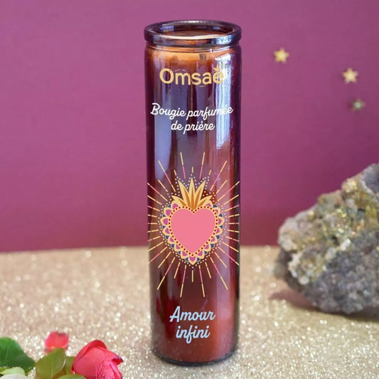 Bougie parfumée de prière Amour infini, Omsae, en verre brun avec un Sacré coeur doré et rose, posée sur du sable, à côté d'une pierre et d'une rose