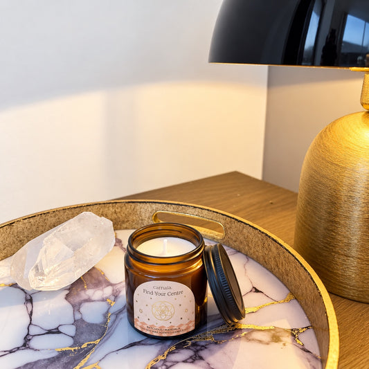 Bougie Carnatia Find Your Centre ouverte avec cire blanche et couvercle noir posé à côté, accompagnée d’une pierre cristal de roche sur un plateau décoratif marbré près d’une lampe dorée, De A à KA