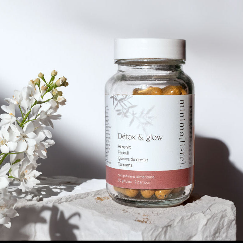 Pot en verre de gélules Detox & Glow avec étiquette minimaliste, posé sur un socle de pierres blanches, entouré de fleurs de cerisier, dans une ambiance pure et lumineuse