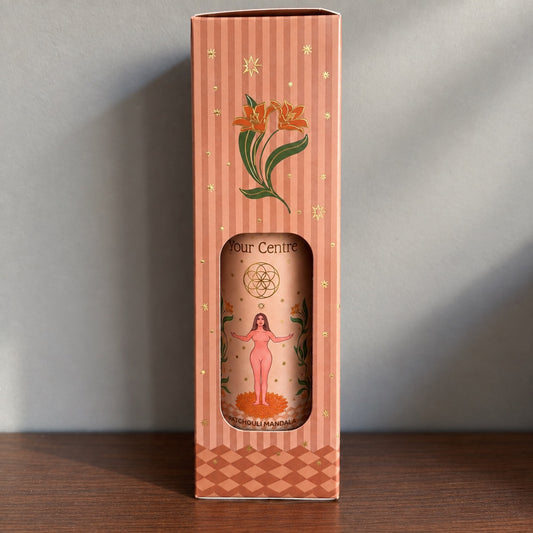 Étui rayé terracota du spray d'ambiance Find Your Centre Carnatia, orné de fleurs dorées et d'étoiles, avec fenêtre découpée laissant apparaître le flacon patchouli mandala et son illustration de femme les bras ouverts.