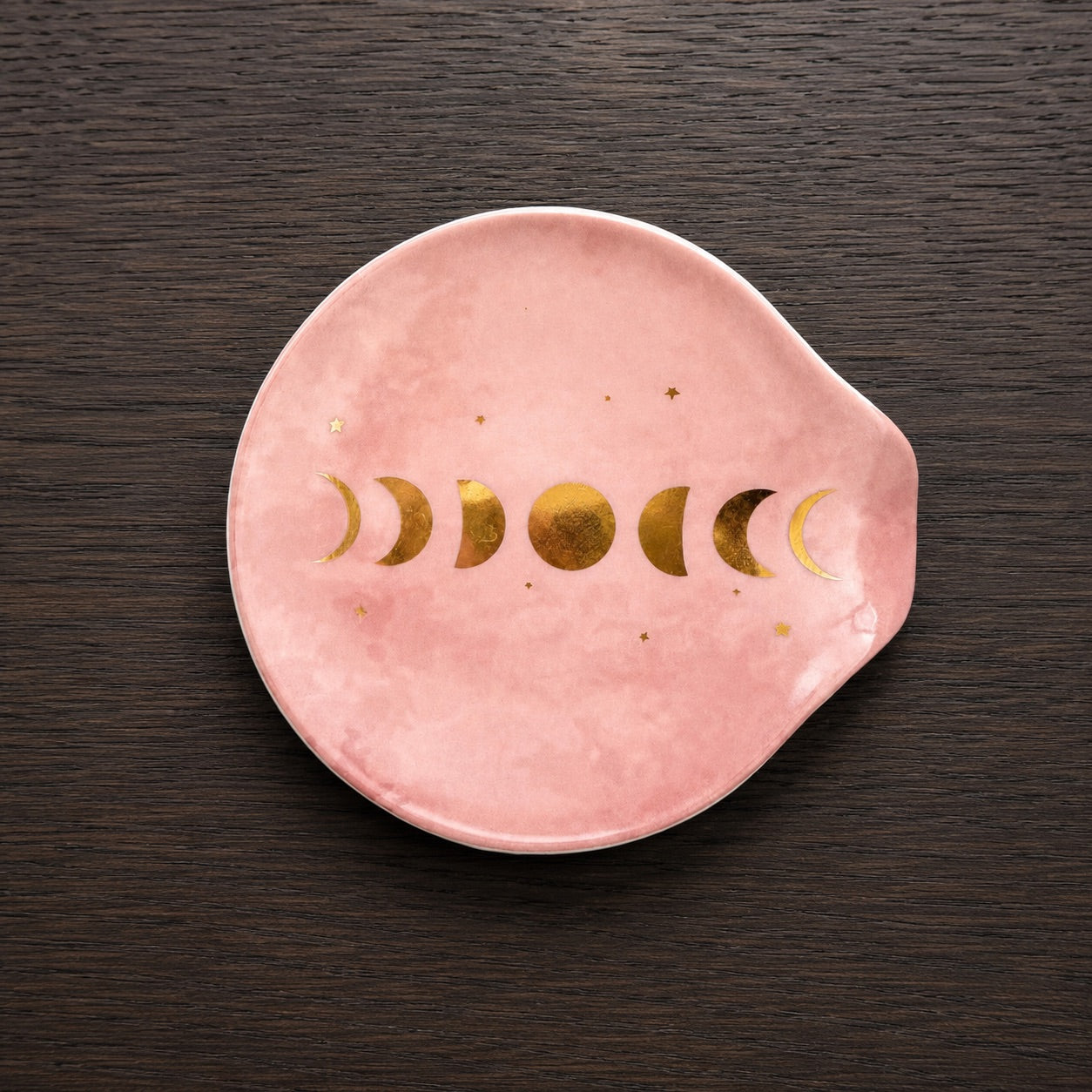 Coupelle porte sachet de thé en porcelaine rose poudrée avec motif phases de la Lune dorées et étoiles, vue de dessus sur fond bois