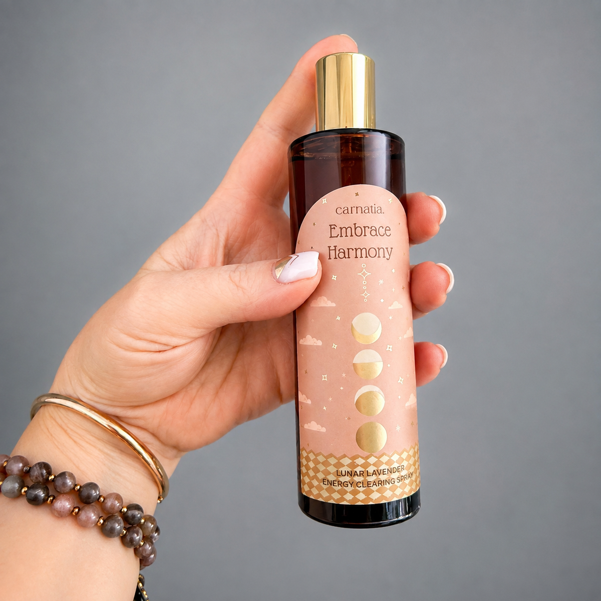 Spray d'ambiance Embrace Harmony Lunar Lavender Carnatia, flacon verre ambré et doré tenu en main, étiquette rose avec phases de lune dorées