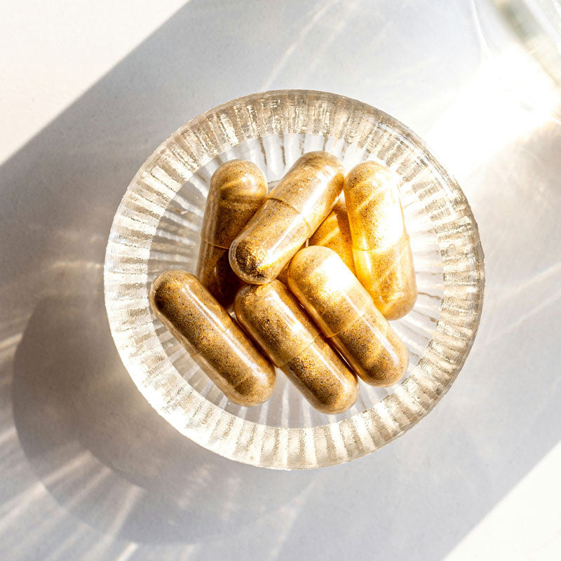 Gélules beiges de compléments alimentaires posées sur une assiette en verre transparent laissant passer les rayons de soleil
