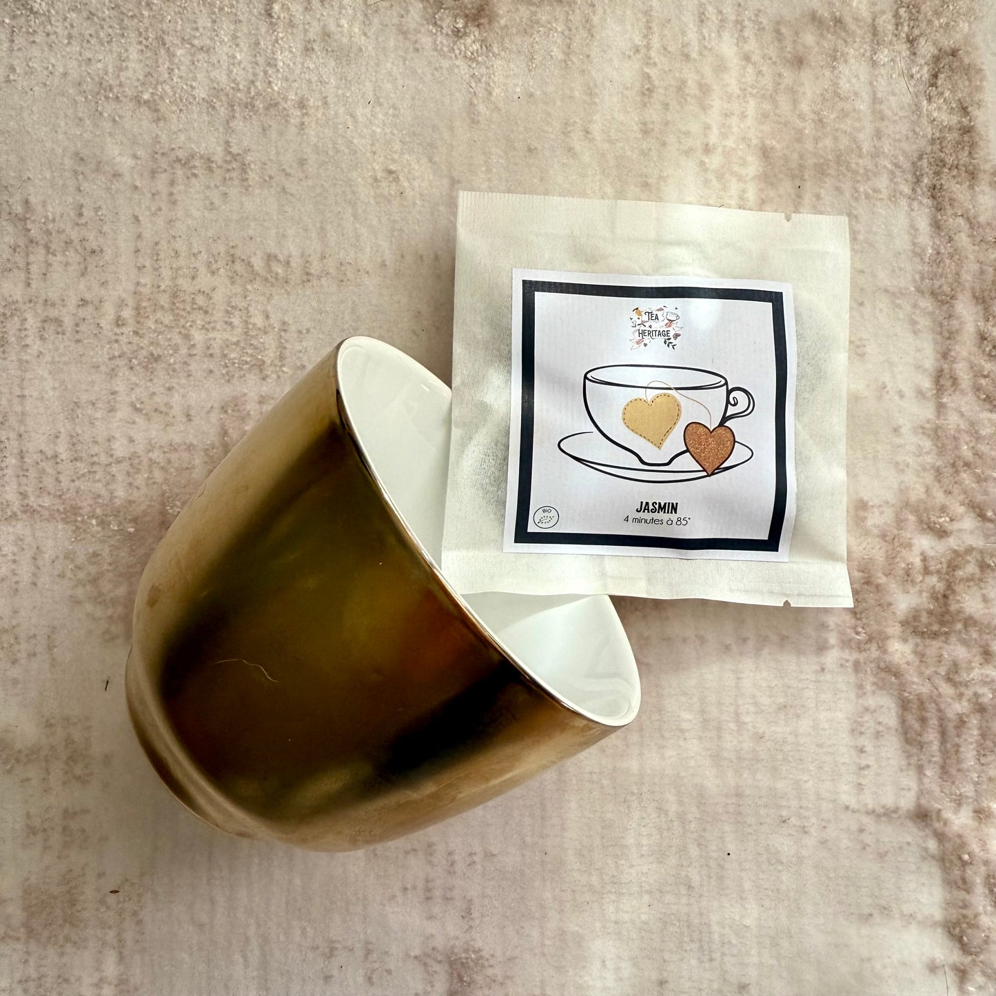 Photo d'ambiance zen avec une tasse de thé dorée couchée sur un tapis ivoire et une pochette de sachets de thé vert au jasmin, avec étiquette de la marque Tea Heritage