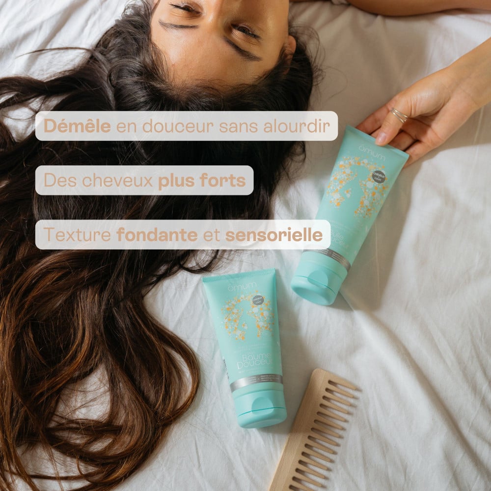 Visuel de l'après shampoing Mon baume douceur d'Omum avec une femme couchée sur un lit tenant un tube turquoise, à côté d'un second tube et d'un peigne en bois