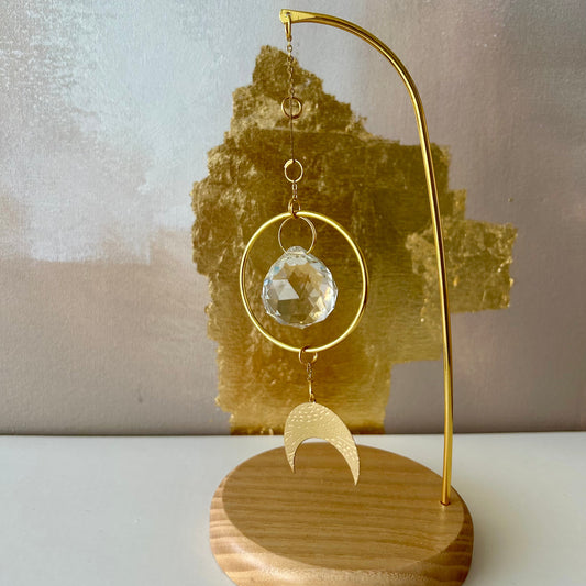 Attrape-soleil Luna Omsaé en laiton et cristal facetté sur un socle en bois de frêne, posé sur uneétagère blanche, décor mural beige et feuilles d'or, Feng Shui