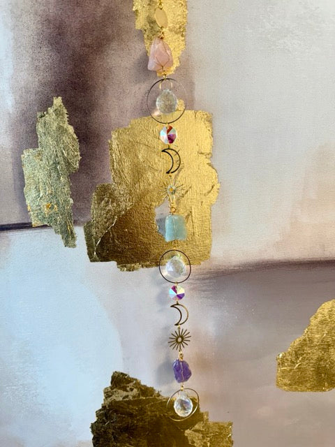 Attrape soleil suspendu devant une toile or, pierres naturelles, cristal de roche, symbole de lune et soleil en métal doré