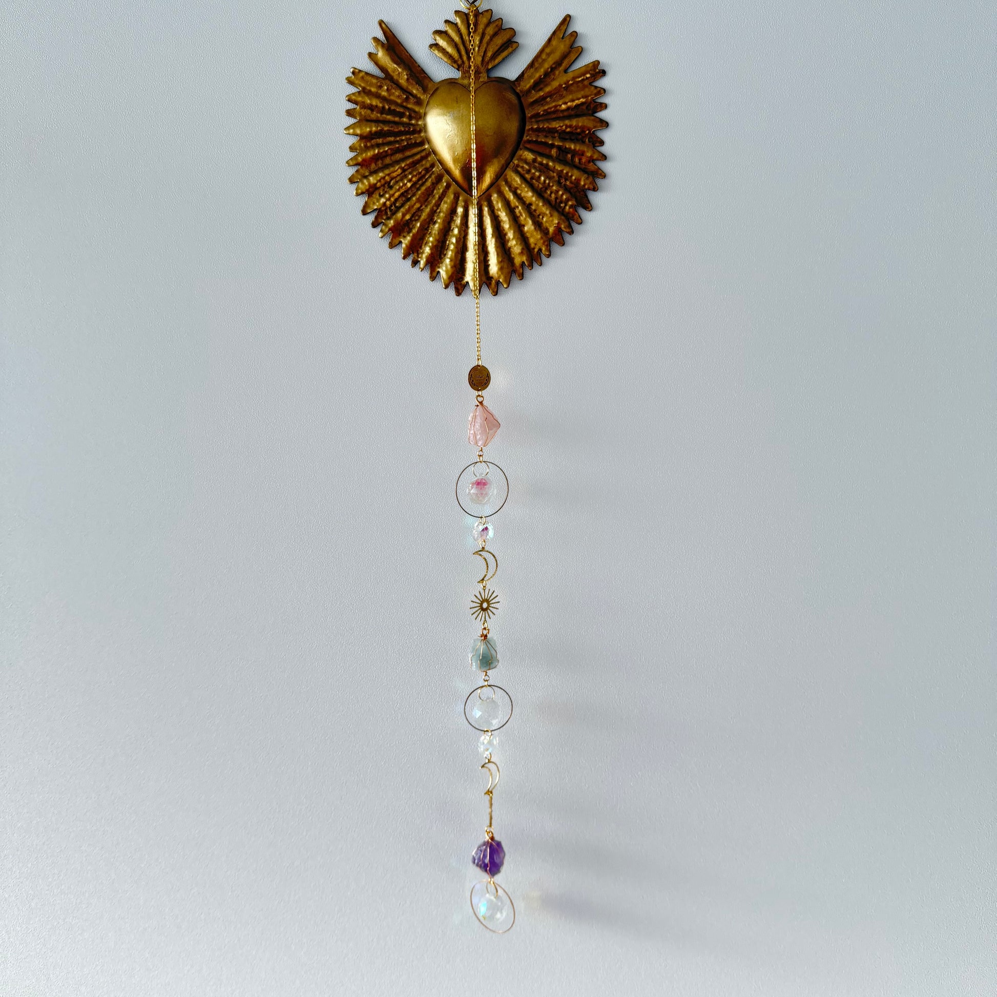 Sacré coeur or accroché sur un mur blanc avec attrape soleil doré, cristal de roche, quartz rose, améthyste, fluorite, lune et soleil