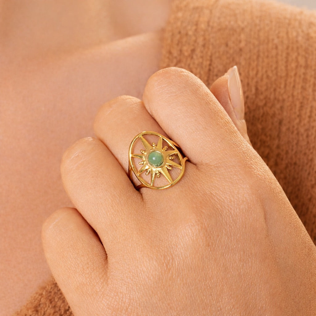 Main de femme pliée avec bague étoile dorée et pierre ronde d'aventurine verte au centre, contre pullover camel