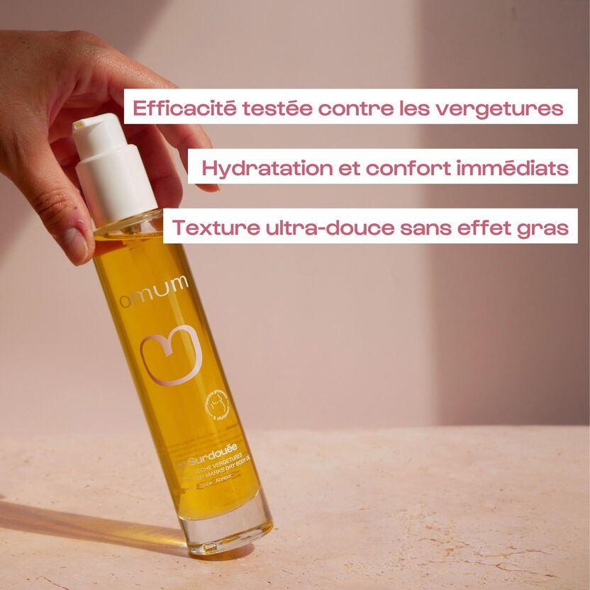 Main tenant un flacon pompe de l'Huile corps anti vergetures La Surdouée d'Omum avec ses bienfaits, efficacité et texture, fond rose beige clair
