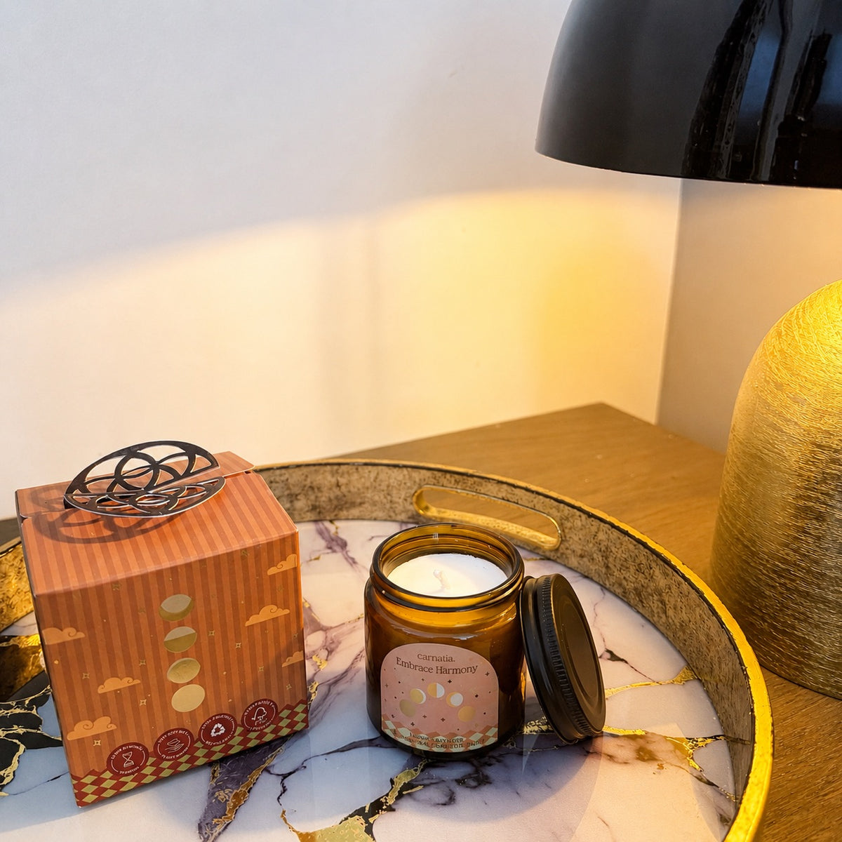 Bougie parfumée Lunar Lavender Carnatia dans un pot en verre ambré ouvert avec son couvercle noir posé à côté et son emballage orange sur un plateau décoratif dans une ambiance chaleureuse, De A à KA