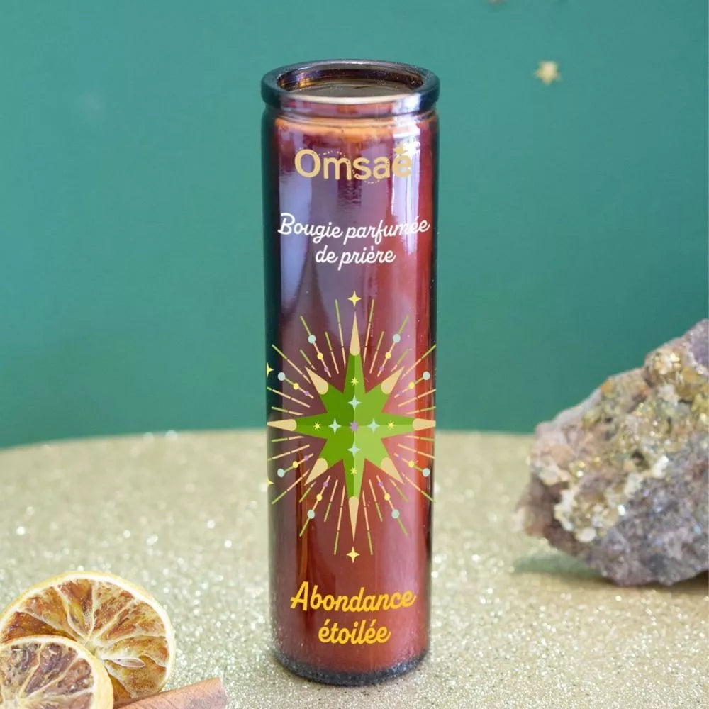 Bougie parfumée de prière Abondance étoilée, Omsae, en verre brun, avec étoile dorée et verte au centre, posée sur du sable, tranches d'oranges séchées et pierre