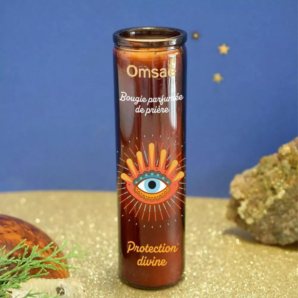 Bougie parfumée de prière Protection divine Omsae, en verre brun avec un oeil bleu et doré, posée sur du sable, à côté d'une pierre 