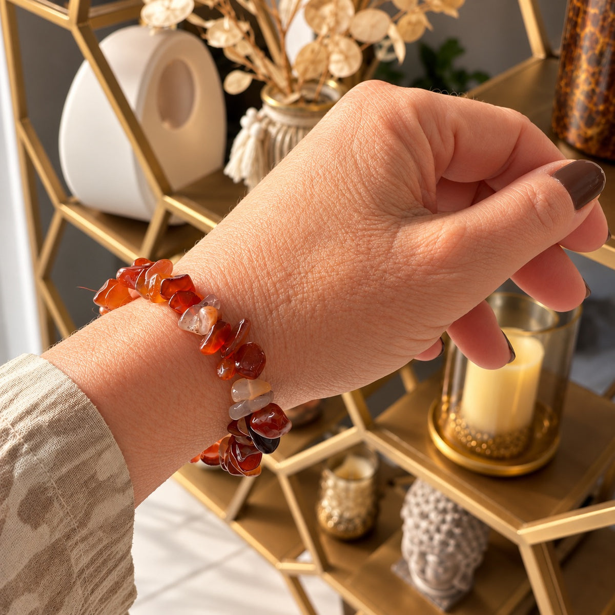 Bracelet en pierres de cornaline aux nuances orangées et rouges, porté au poignet, devant une étagère dorée avec une bougie et des objets décoratifs
