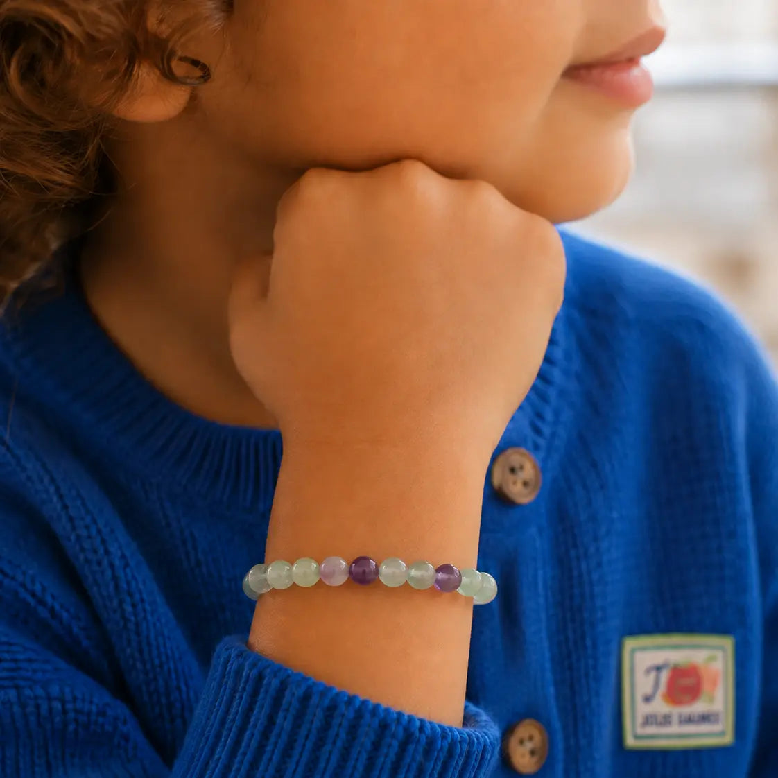 Bracelet enfant en perles de fluorite vert pâle et violettes porté au poignet avec un gilet bleu
