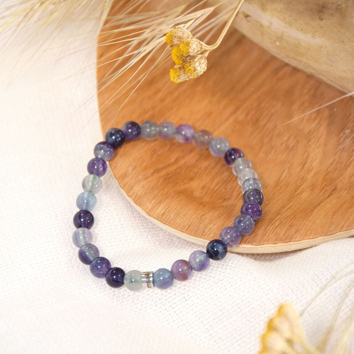 Bracelet Omsaé en perles rondes de fluorite mauve, violet et perle en acier argenté, posé sur une plaque de bois et tissu blanc avec fleur séchée jaune