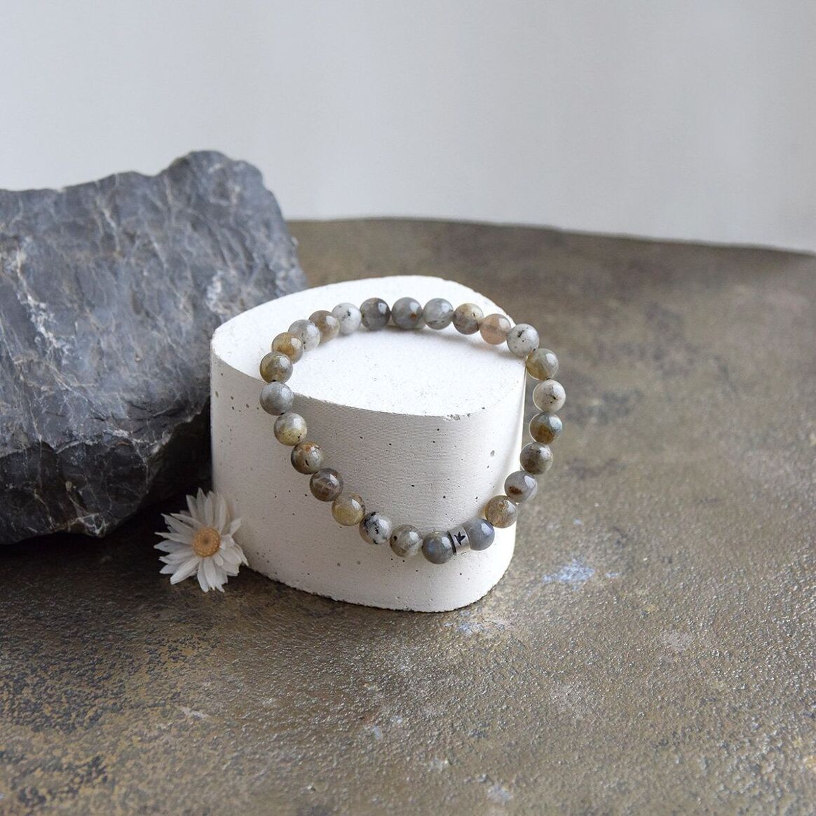 Bracelet en perles naturelles de labradorite grise, posé sur un sockle blanc, avec une fleur et une grosse pierre