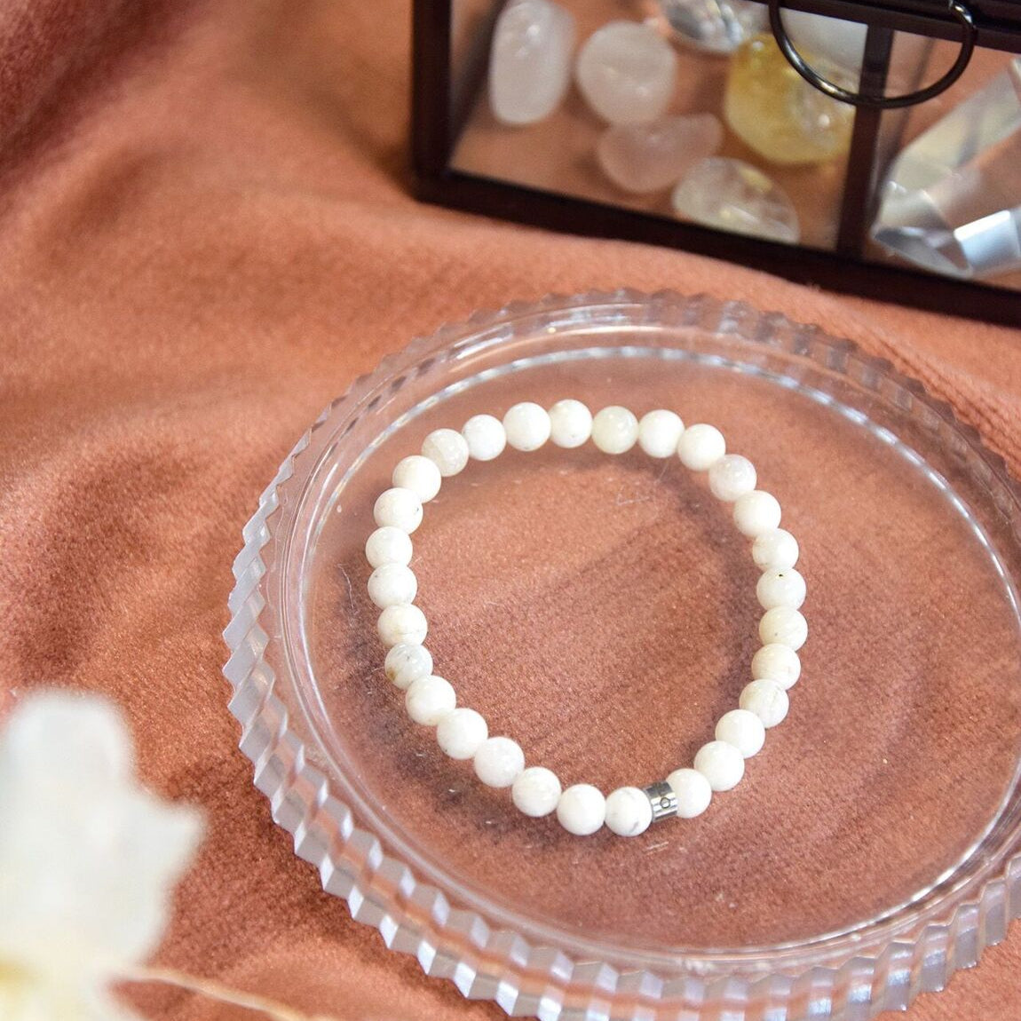 Bracelet en perles de pierre de lune blanche posé sur une coupelle en verre à côté d'une boite en verre contenant des cristaux