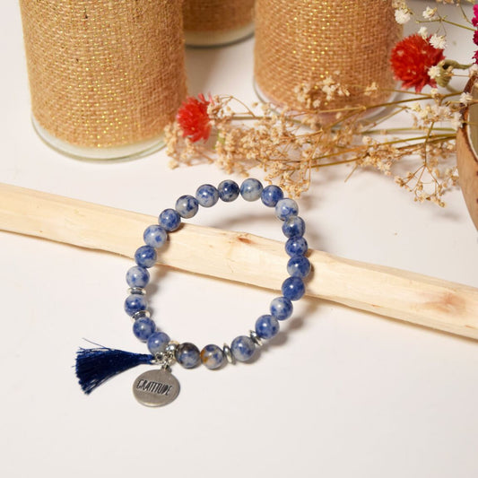 bracelet en perles naturelles de sodalite bleu, avec pompon en coton et charm gratitude en métal argenté, posé sur du bois, à côté de fleurs séchées et de bocaux