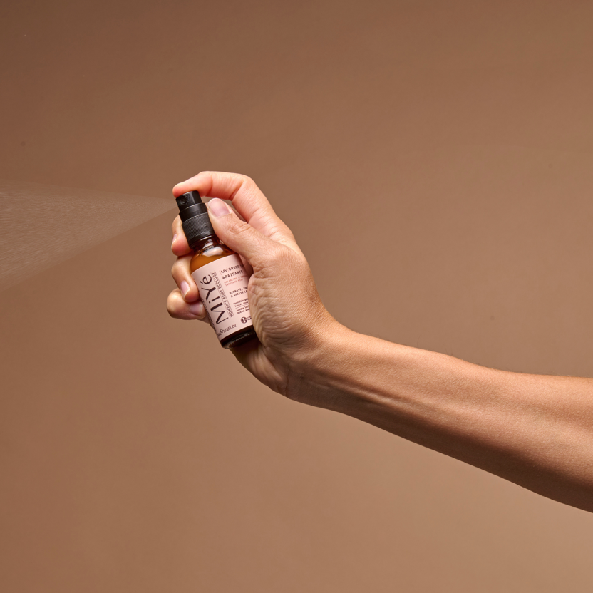 Main de femme vaporisant la brume intime apaisant MiYé, flacon verre brun, étiquette rose, fond ocre