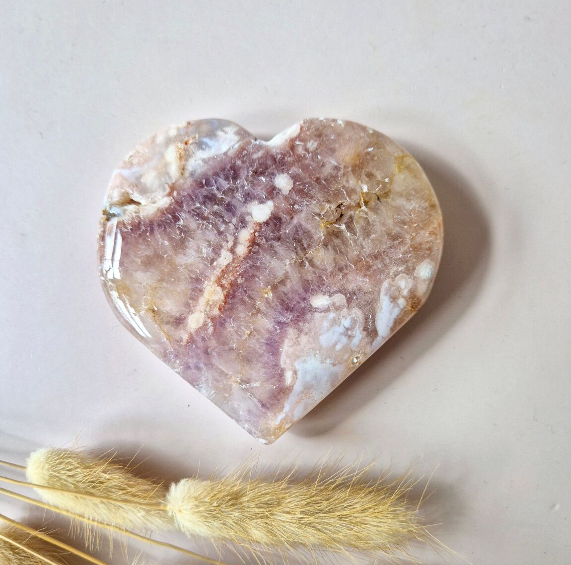 Coeur en améthyste rose drusée, posée sur un fond rose clair, accompagné de brins de pampas séchées