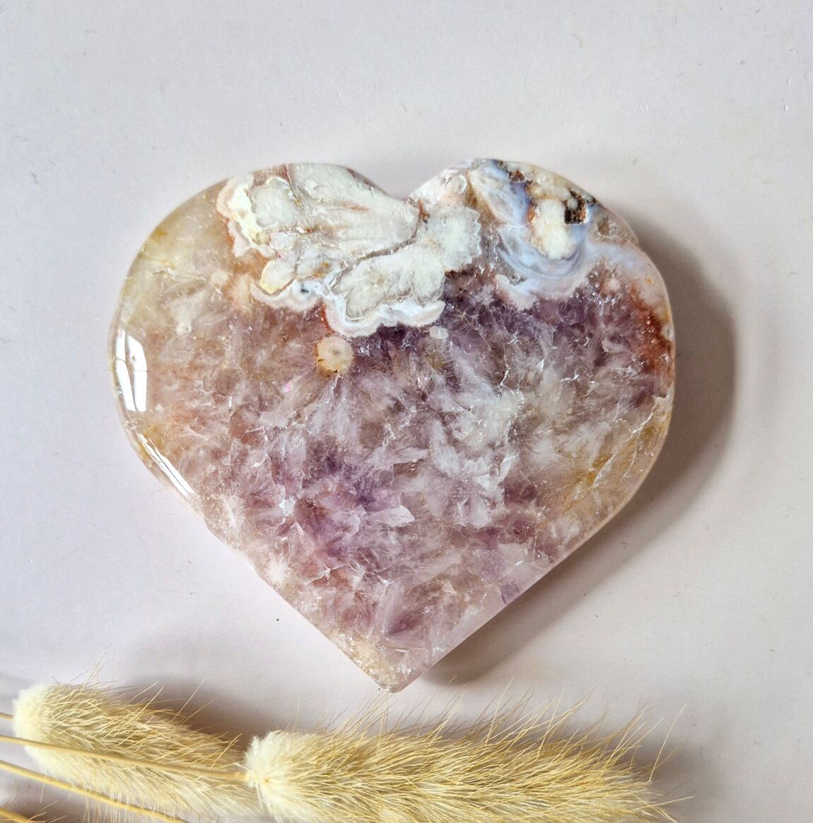 Coeur en améthyste rose drusée, posée sur un fond rose clair, accompagné de brins de pampas séchées, pierre apaisante