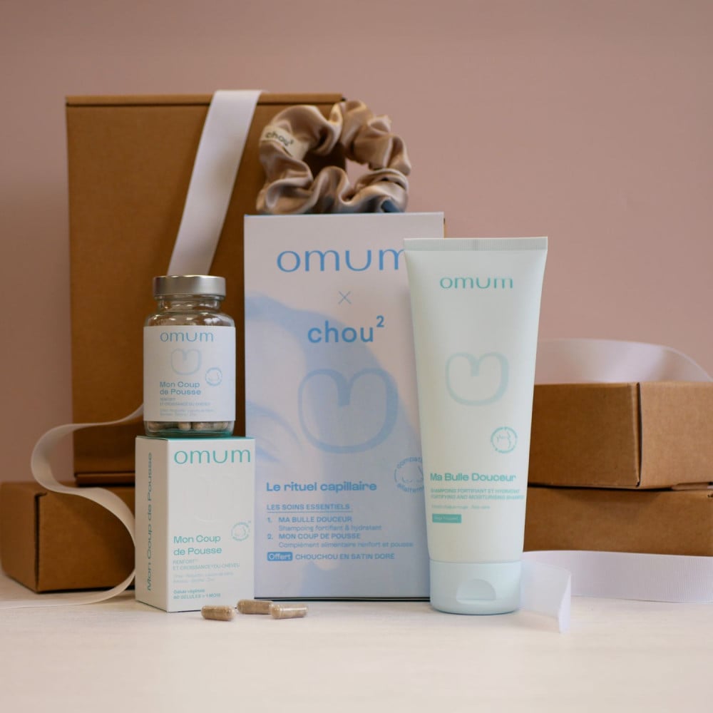 Coffret Omum rituel capillaire composé d'un pot de compléments alimentaires posé sur sa boîte, 3 gélules, d'un tube de shampoing , d'un chouchou doré en satin, sur fond de boîtes en carton