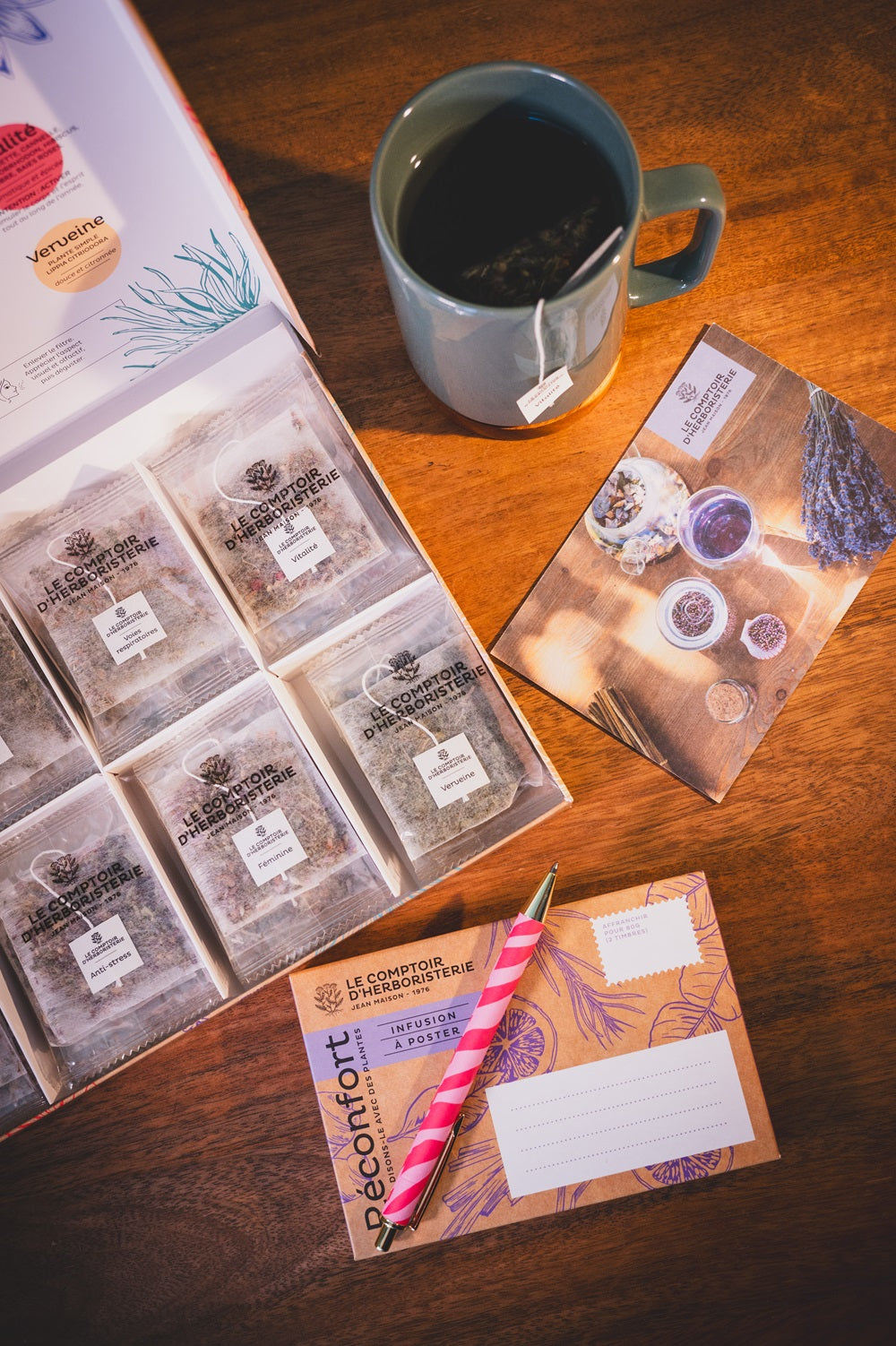 Photo d'ambiance zen avec un coffret ouvert avec des infusions Le Comptoir de l'Herboristerie, une tasse avec une tisane en train d'infuser, une carte postale et une enveloppe avec un stylo
