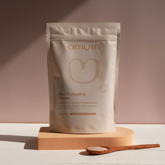 Sachet du complément alimentaire Mon collagène cacao d'Omum, posé sur un socle bois avec une cuillère doseuse bois avec la poudre de collagène sur fond terracota zen