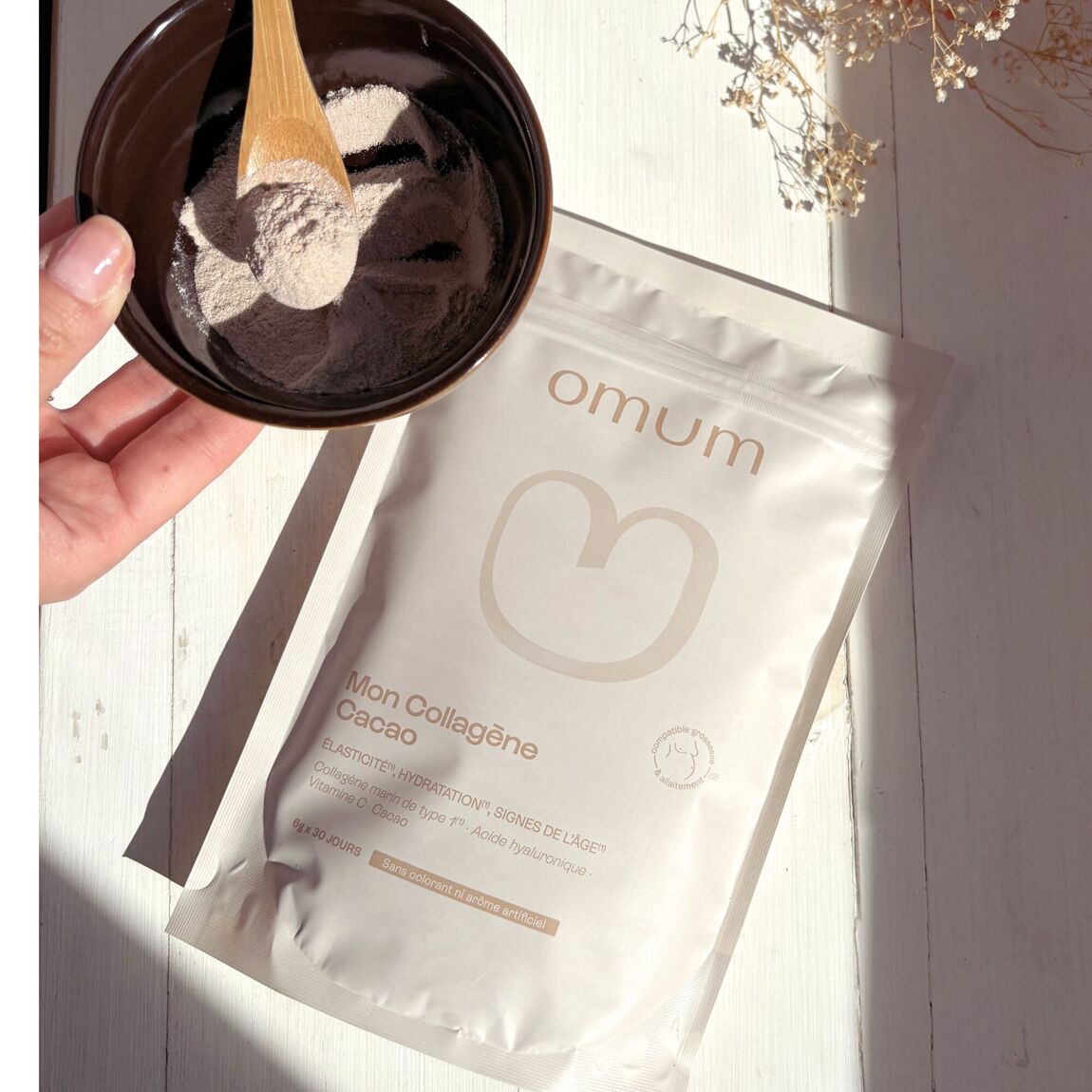 Sachet du complément alimentaire Mon collagène cacao d'Omum avec main tenant un bol avec la poudre cacaotée et une cuillère bois