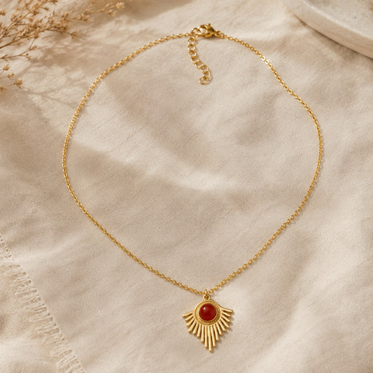 Collier solaire doré avec pierre de cornaline rouge posé à plat sur tissu en lin beige avec fleurs séchées en arrière-plan
