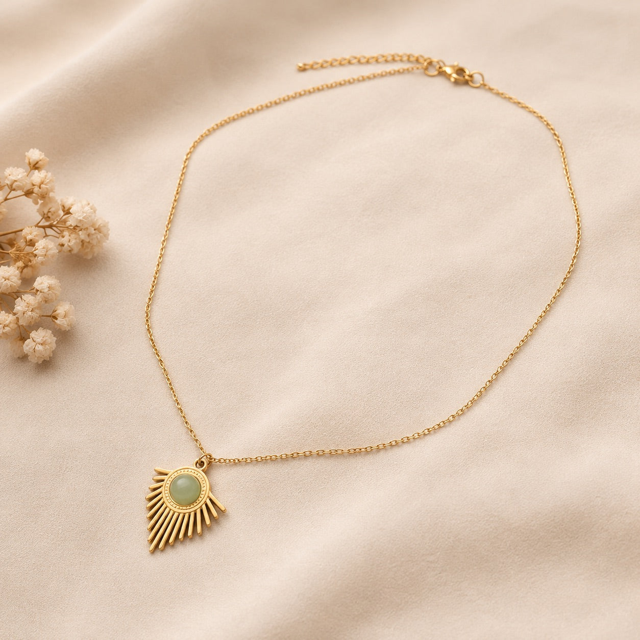 Collier solaire doré avec pendentif et pierre d'aventurine verte posé sur tissu beige, accompagné de fleurs séchées blanches
