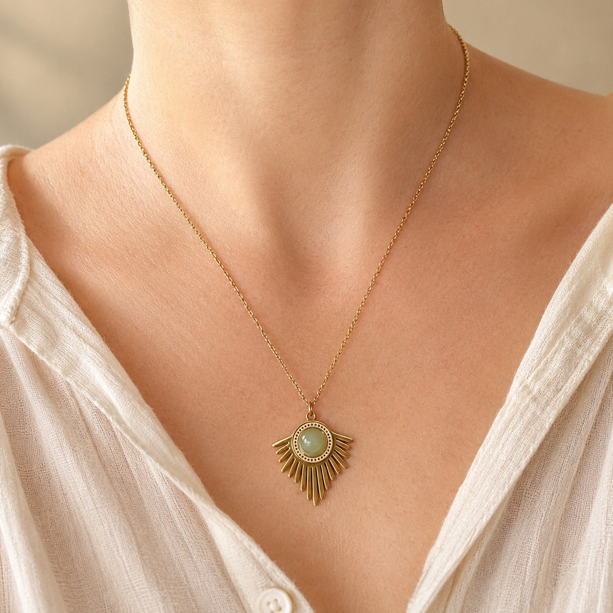 Collier doré avec pendentif solaire et pierre d'aventurine verte porté sur une femme en blouse blanche, fond beige naturel