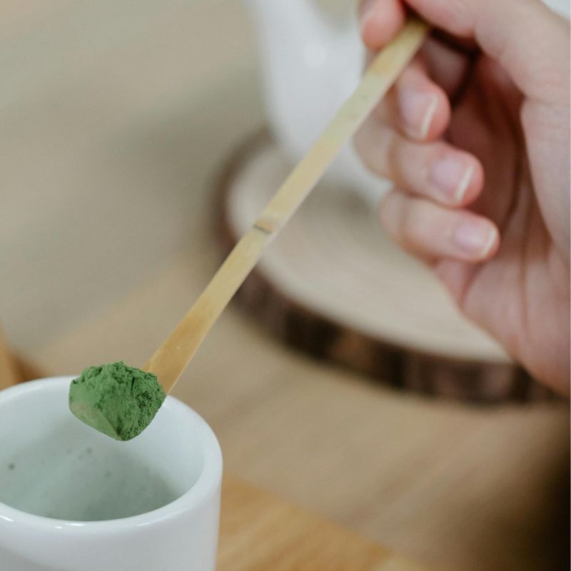 Main tenant un chashaku, cuillère doseuse pour matcha en bambou , poudre verte de matcha au dessus d'une tasse blanche