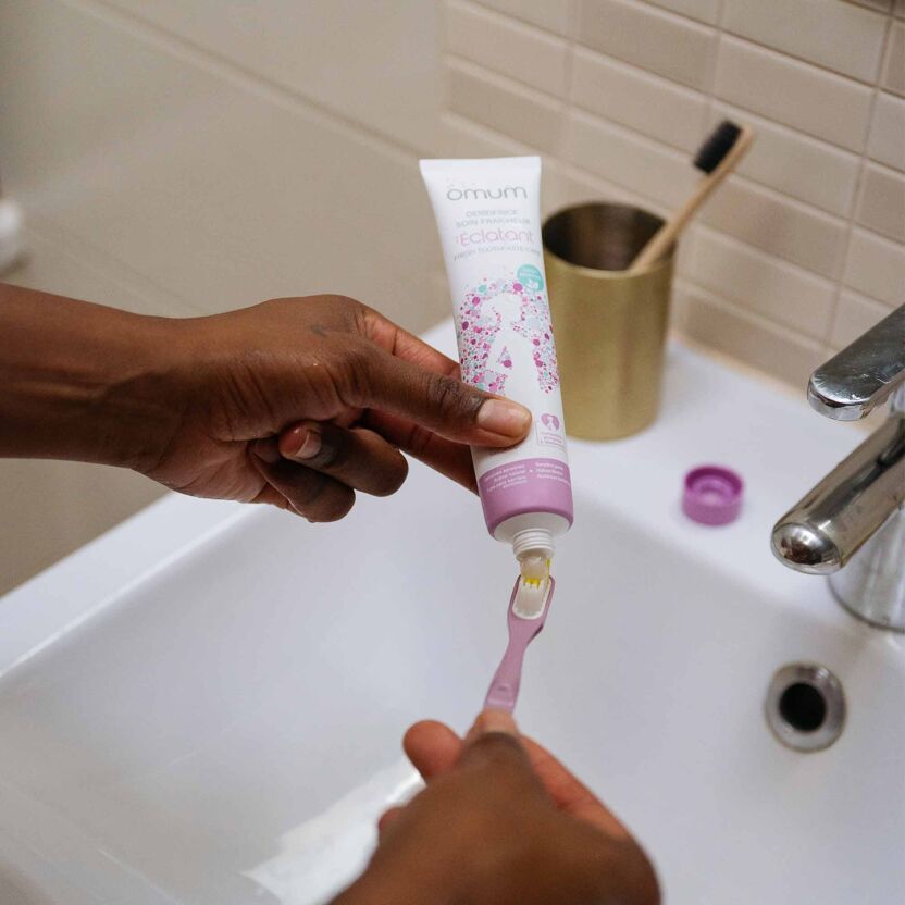 Mains de femmes tenant un tube de dentifrice blanc et violet, l'Éclatant, soin fraîcheur Omum, et l'appliquant sur une brosse à dents violette, lavabo blanc