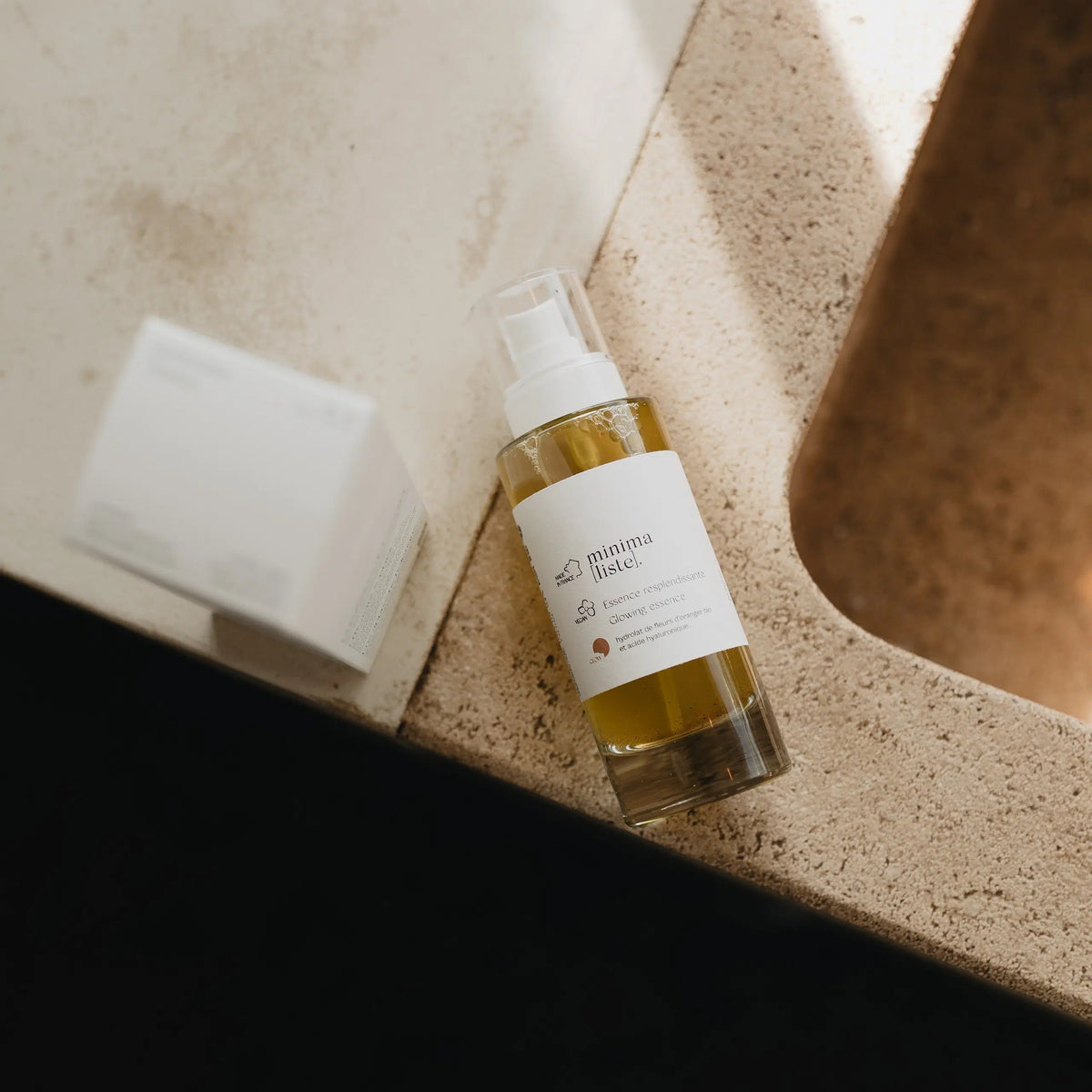 Flacon verre d’Essence Resplendissante Minima[liste] posé sur un lavabo en pierre claire, à côté de son étui blanc, dans une lumière dorée