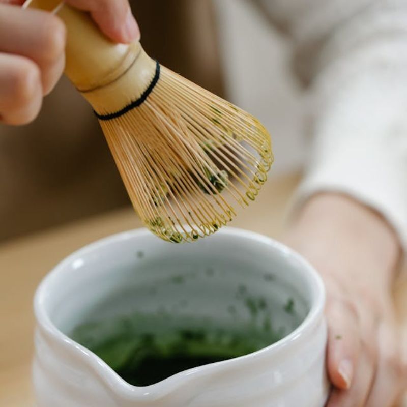 Main tenant un fouet matcha en bambou, chasen, pour mélanger son thé vert matcha dans un bol en porcelaine blanche avec bec verseur