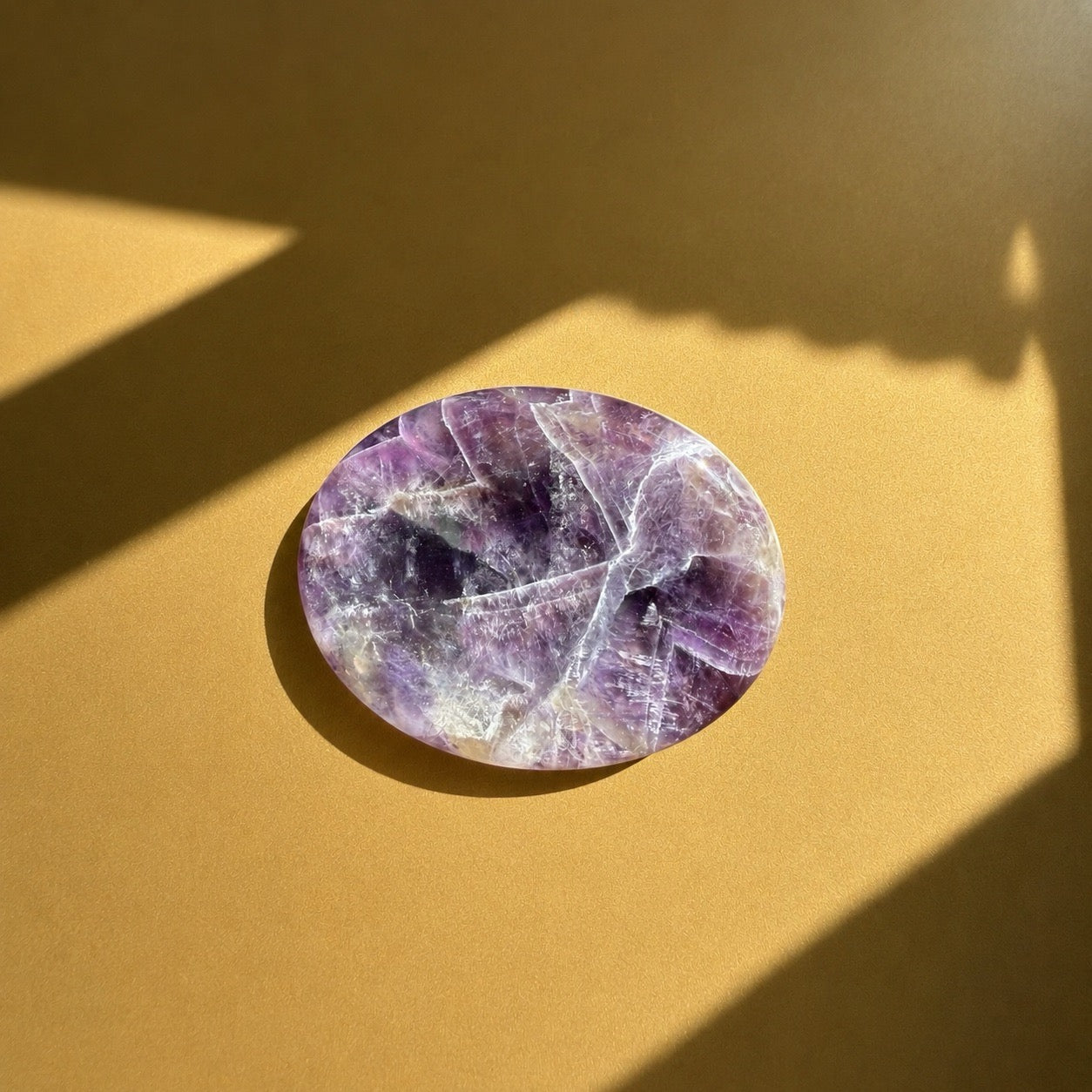 Galet anti stress plat en améthyste polie aux effets violets et blancs, posé dans une lumière dorée avec jeux d'ombre, De A à KA