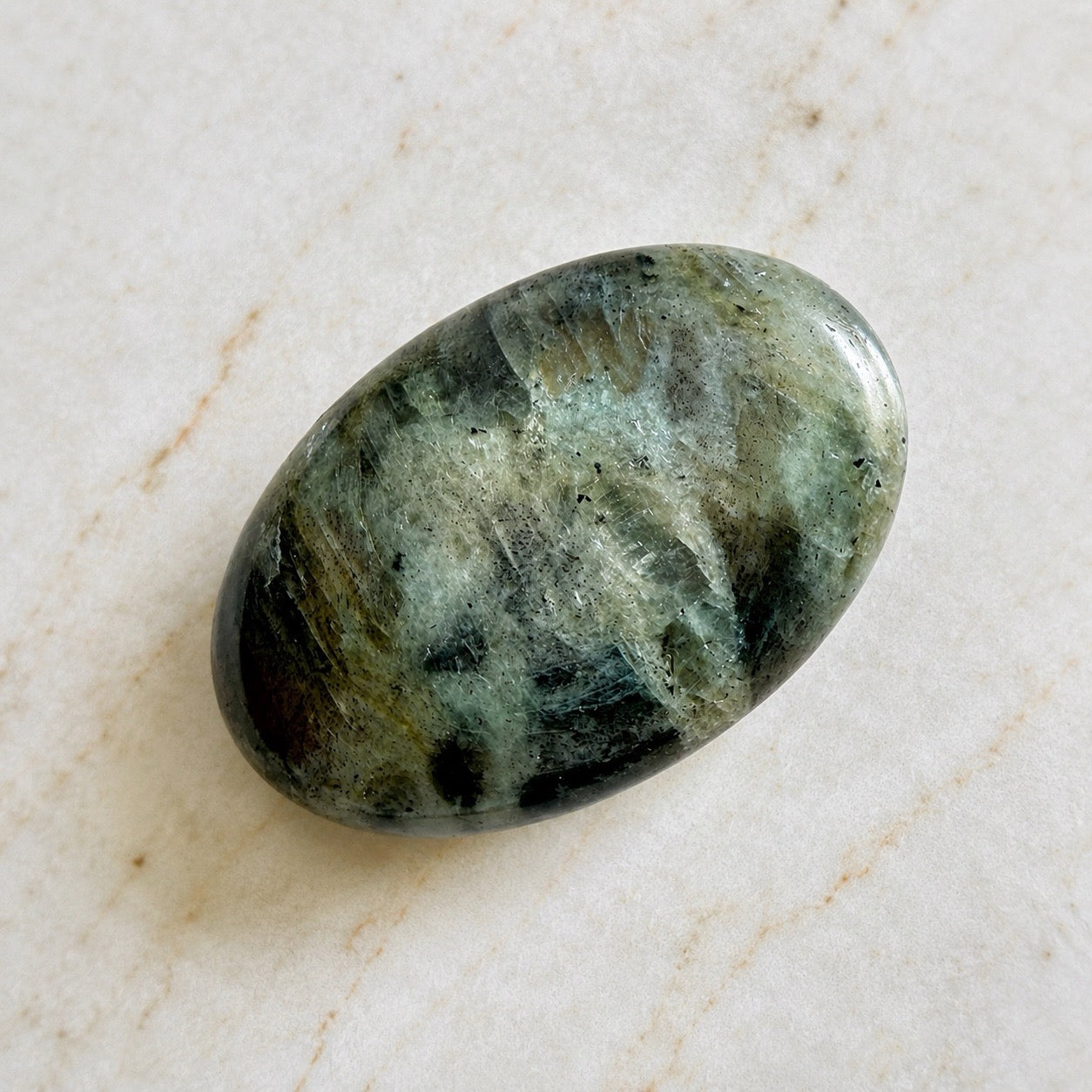 Galet de pierre naturelle de labradorite gris aux reflet verts et dorés, sur table en marbre clair beige, protection et intuition
