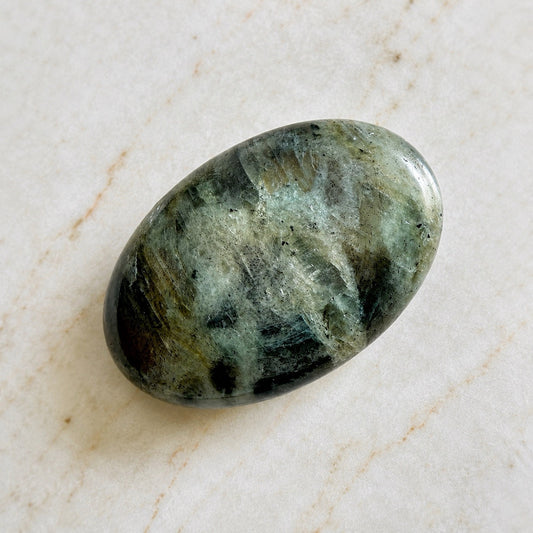 Galet de pierre naturelle de labradorite gris aux reflet verts et dorés, sur table en marbre clair beige, protection et intuition
