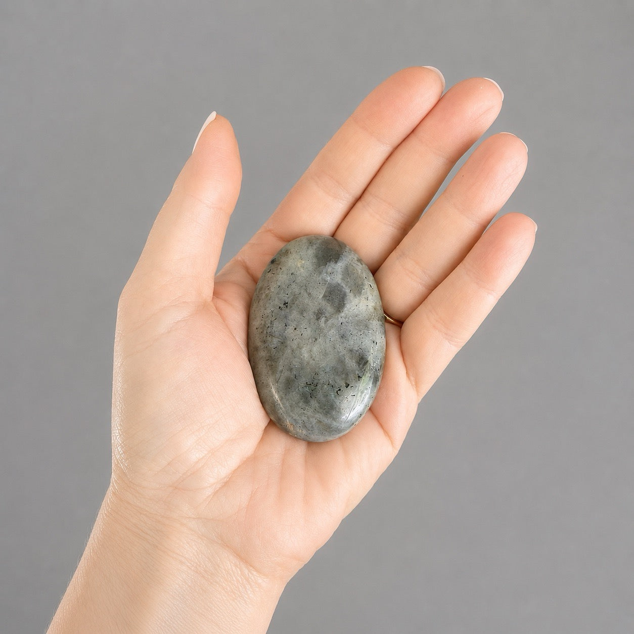 Main tenant un galet de pierre naturelle de labradorite gris, vert, sur fond gris, protection énergétique et intuition en lithothérapie