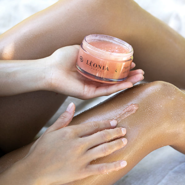 Application du gommage corps bio Thé Blanc Lacté Léonia Paris sur les jambes , texture exfoliante douce et nourrissante, orangée rose
