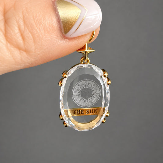 Grigri pendentif The Sun en cristal de roche facetté tenu entre les doigts, monture en cuivre plaqué or avec inscription The Sun et motif soleil gravé
