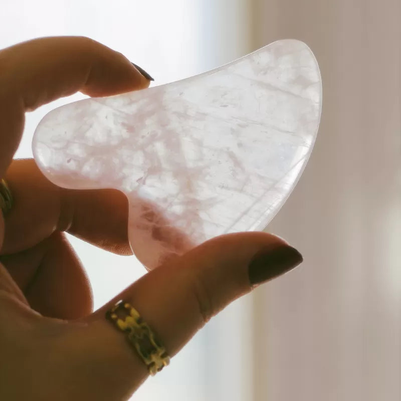 Gua sha en quartz rose tenu entre les doigts devant une lumière douce