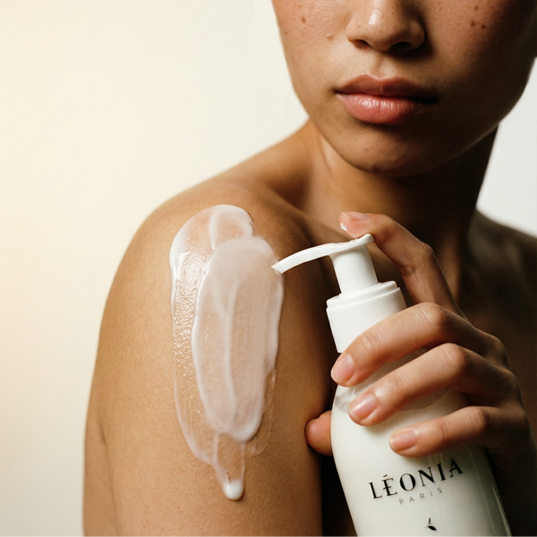 Personne appliquant du lait pour le corps sur son épaule avec un flacon pompe blanc Léonia Paris à la main