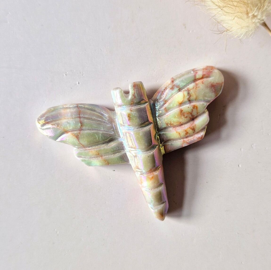 Libellule en quartz angel aura posée sur un fond beige avec herbe séchée, pierre énergétique irisée symbole de liberté et de transformation, boutique De A à KÂ
