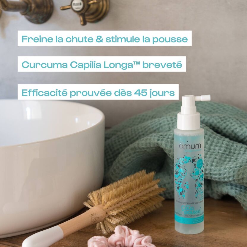 Visuel de la lotion fortifiante anti-chute de cheveux Omum avec ses bienfaits poto du produit sur un meuble de salle de bain en bois avec chouchou rose, brosse en bois et serviette verte